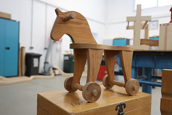 Holzspielzeug in Form eines Pferdes auf vier Rädern, stehend auf einem Holzkasten in einer Werkstatt.