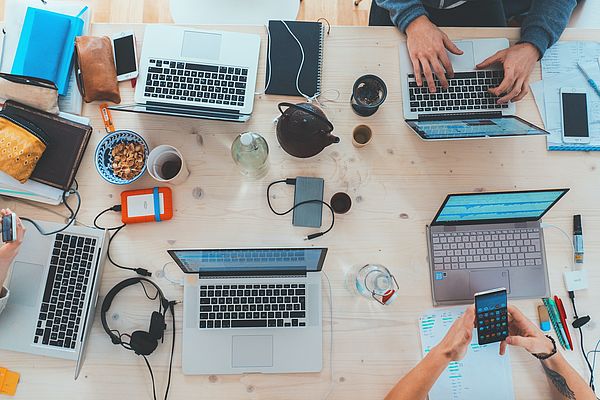 Mehrere Laptops, Kopfhörer, Handys, Notizbücher und Getränke auf einem Holztisch, Menschen arbeiten gemeinsam an einem Projekt.