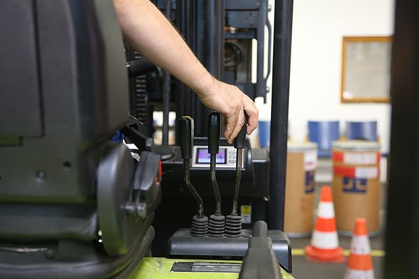 Hand bedient Hebel an einem Gabelstapler in einem Lager mit Kartons und Verkehrskegeln im Hintergrund.