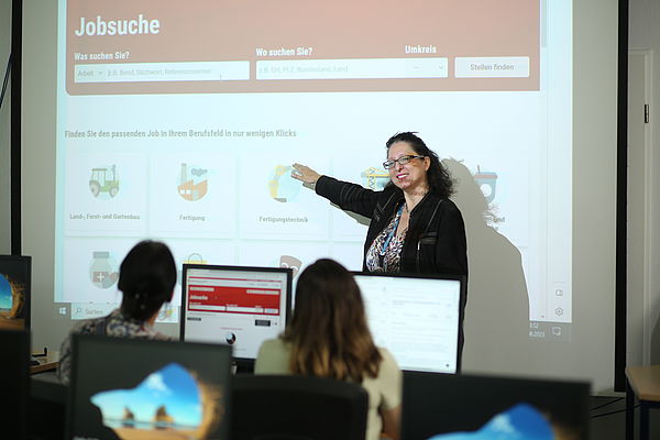 Frau präsentiert eine Jobsuche-Webseite vor Publikum in einem Klassenraum mit Computern.