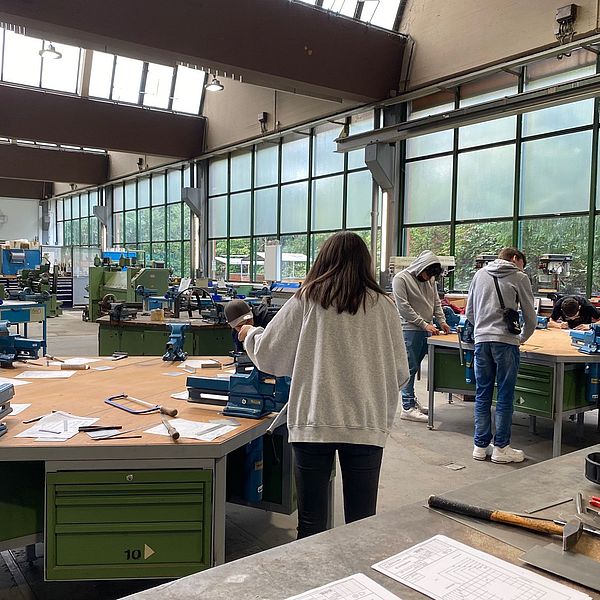 Junge Menschen arbeiten an Werkbänken in einer hellen Werkstatt mit großen Fenstern und Maschinen im Hintergrund.