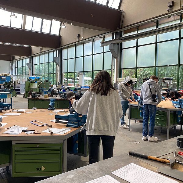 Junge Menschen arbeiten an Werkbänken in einer hellen Werkstatt mit großen Fenstern und Maschinen im Hintergrund.