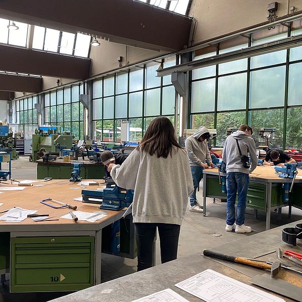 Junge Menschen arbeiten an Werkbänken in einer hellen Werkstatt mit großen Fenstern und Maschinen im Hintergrund.