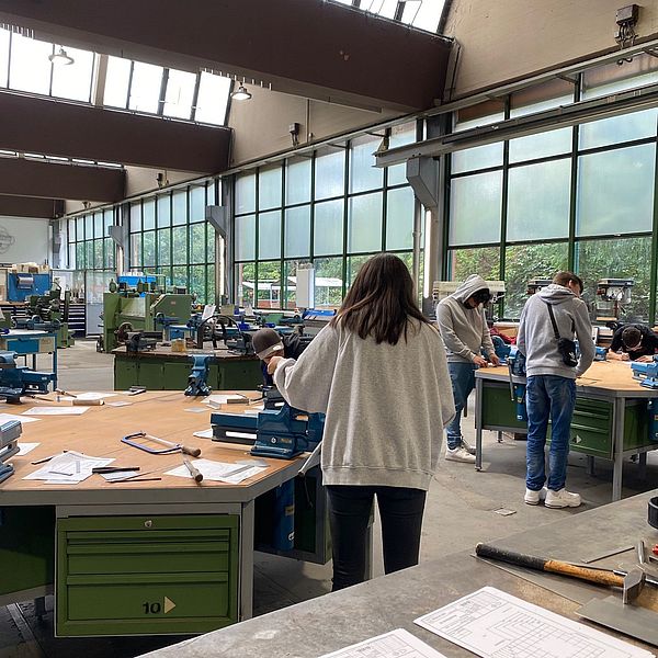 Junge Menschen arbeiten an Werkbänken in einer hellen Werkstatt mit großen Fenstern und Maschinen im Hintergrund.