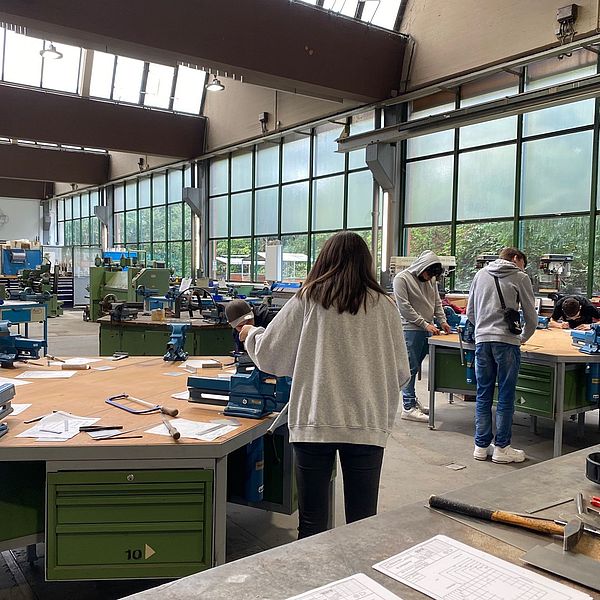 Junge Menschen arbeiten an Werkbänken in einer hellen Werkstatt mit großen Fenstern und Maschinen im Hintergrund.
