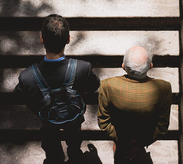 Zwei Männer, einer älter mit grauem Haar und einer jünger mit dunklem Haar, laufen nebeneinander auf einer Treppe mit Schatten und Lichtstreifen.