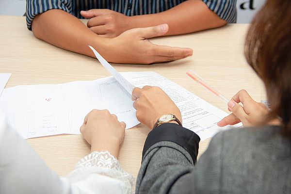 Drei Personen sitzen an einem Tisch und besprechen Dokumente, eine Person hält ein Blatt Papier in der Hand.