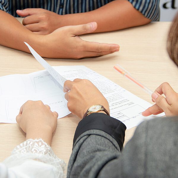 Drei Personen sitzen an einem Tisch und besprechen Dokumente, eine Person hält ein Blatt Papier in der Hand.