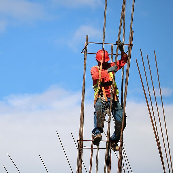 Bauarbeiter mit rotem Helm und Sicherheitsweste klettert an Stahlarmierungen auf einer Baustelle vor blauem Himmel.