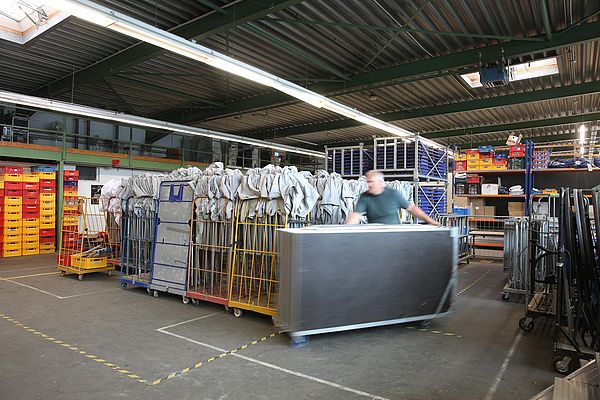 Lagerhalle mit Regalen und Transportwagen, eine Person bewegt einen großen Metallbehälter.