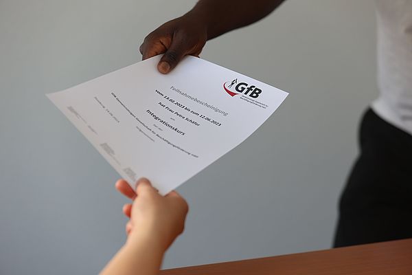 Zwei Personen tauschen ein Dokument mit dem Logo der GfB aus, das eine Teilnahmebestätigung enthält.