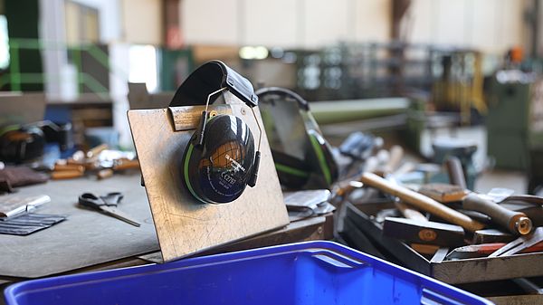 Gehörschutz auf Metallplatte in einer Werkstatt mit Werkzeugen und Maschinen im Hintergrund.