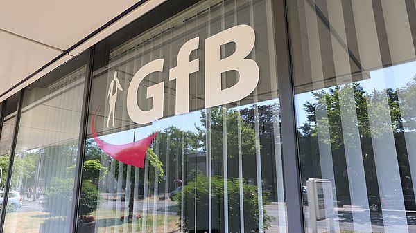 Glasfassade mit weißem GfB-Logo, reflektiert Bäume und Straße.
