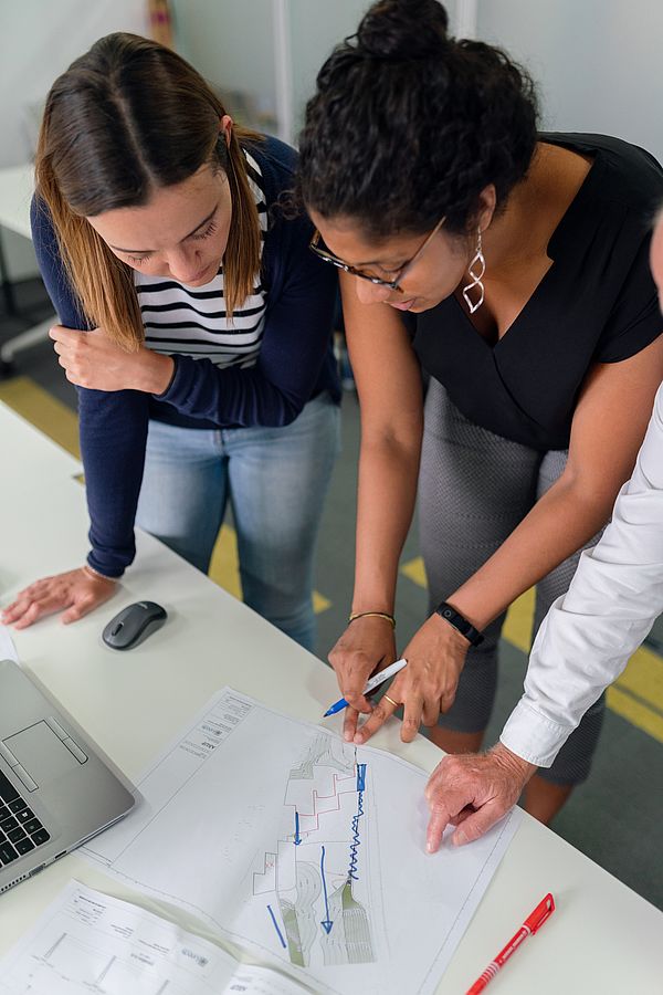 Drei Personen diskutieren und zeigen auf ein Papier mit Diagrammen auf einem Tisch neben einem Laptop und einer Maus.